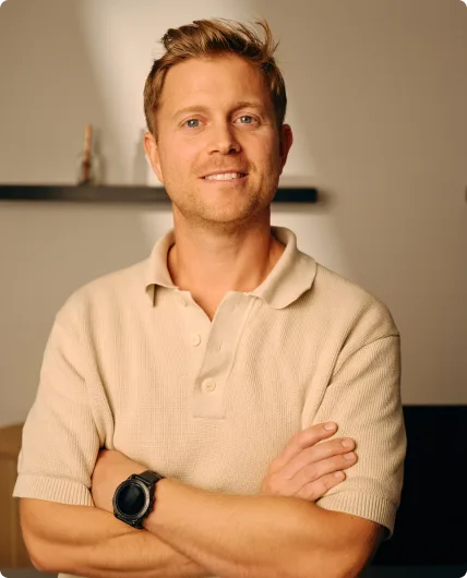 Man with reddish-brown hair and crossed arms wearing beige polo shirt with dark watch